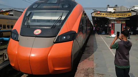 Jamie Fullerton An orange Vande Bharat Express sleeper train stands at a station platform as a boy takes a photo of it (Credit: Jamie Fullerton)
