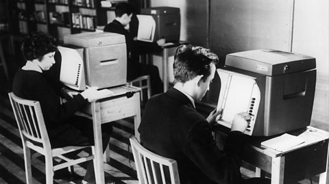 A black and white photo of students learning to use computers in the 1960s. Three students sit in uniforms - white shirts and blazers - on chairs looking at paper graphs with big computer screens in front of them on desks
