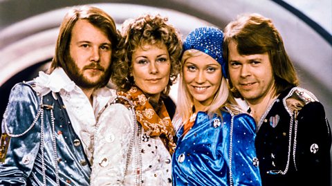 ABBA on stage at Eurovision. One member is in a blue suit, next to the two female singers - one in an orange and white jumpsuit with a skirt and the other in a blue bandana with a blue jumpsuit and on the far right is another male member wearing a long sleeved black outfit