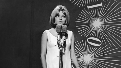A black and white photo of singer France Gall on stage singing into a mic. She has a white dress on and bobbed hair in a middle part