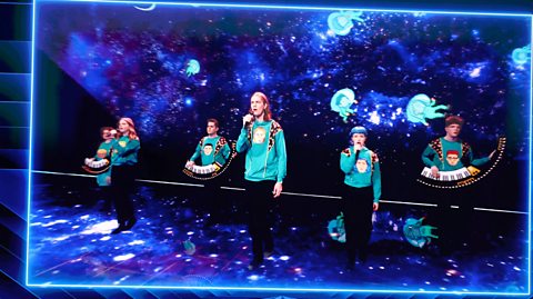 Daði & Gagnamagnið during a pre-recorded performance on the Eurovision stage. All members wear bright blue knitted jumpers with caricatures of themselves knitted at the centre. They all wear black pants and some members hold mics and sing while others have keyboards strapped around their bodies. The stage is full of neon lights and shows a starry sky like in space