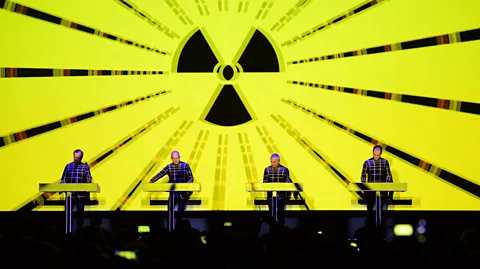 Getty Images The four members of Kraftwerk performing on stage with a radioactive symbol against a bright neon yellow screen behind them (Credit: Getty Images)
