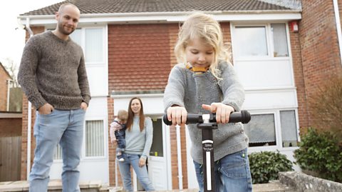 Young girl on scooter watched by father and mother holding toddler in front of typical British semi-detached house