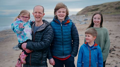 A family of five smiling on a beach, to the left of the photo is Sidney, held up by her carer, next to him his teenage son, and younger son and young daughter.