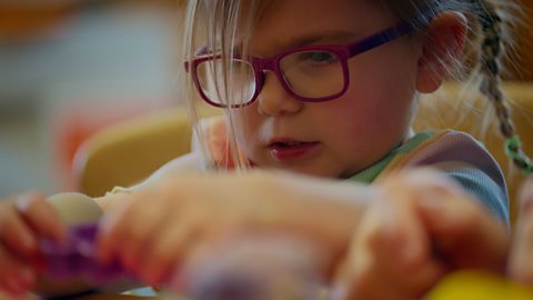 A close-up of a young girl, who is wearing glasses and playing with a toy.