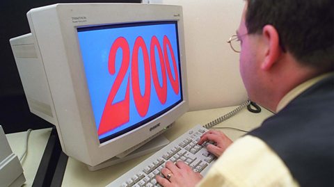 Man sits at a computer typing into the keyboard wearing glasses, a yellow shirt and a grey suit vest. His screen his blue with bold red text reading '2000'