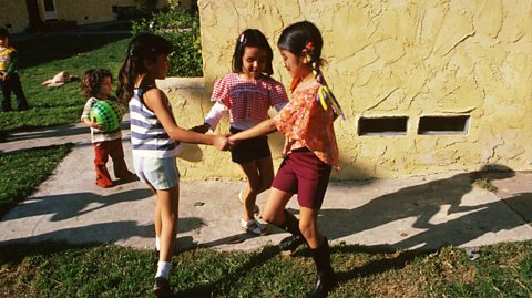 In a sunny garden on the grass, three young girls hold hands in a circle playing ring o'roses. They all smile and wear colourful shorts and tops