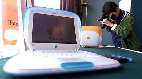 An early 2000s Apple laptop. It is white with a blue shell case and the screen is curved. Behind, a photographer takes a photo of the same laptop but with an orange shell laptop lid