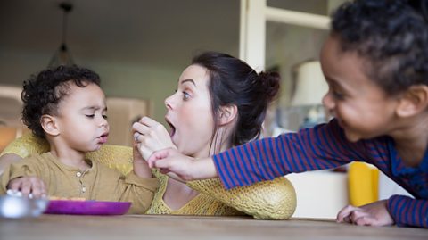 Mum and sibling feed food to a tot at table
