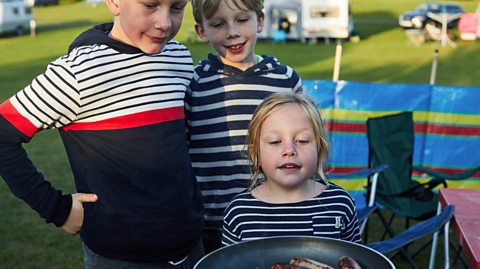 3 young siblings look at a pan of sausages on a campsite