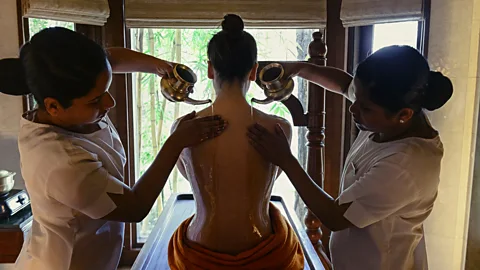 Courtesy of Ananda in the Himalayas Two spa workers pour liquid from bronze beakers onto a woman's back (Credit: Courtesy of Ananda in the Himalayas)