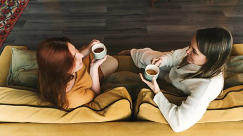 Two female friends sit on a yellow couch smiling and talking while both holding a hot cup of tea