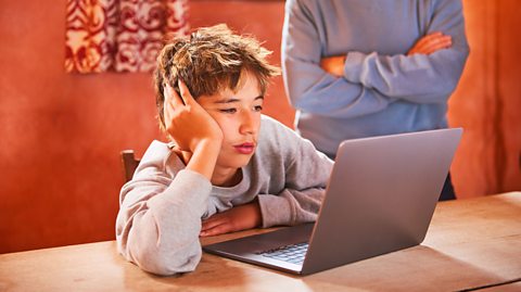 teen boy looks fed up while trying to revise overseen by parent with arms folded