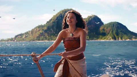 Disney Catherine Laga'aia navigates a sailboat's rudder at sea with green mountains in the distance in Moana (Credit: Disney)