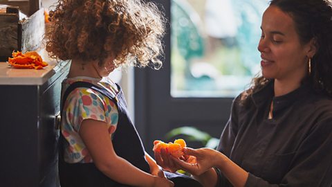 A woman shares some fruit with a small girl in their kitchen.