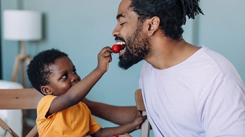 A young boy holds a strawberry while a man takes a bite.