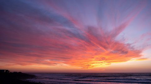 A landscape image of a sunset. The sky features streaks of red, orange, yellow and greyish blue, with red being the most prominent colour. 