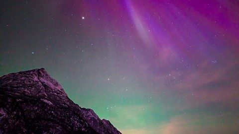 A landscape image that shows silhouetted mountains in the foreground. The sky behind them shows the Northern Lights, with glowing green, pink and purple lights. 