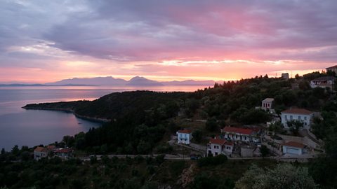 A landscape image of a sunrise over an island. The sky is in pale blues, pinks and oranges, with the sea and the island in the foreground.