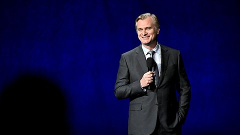 An image of director Christopher Nolan. He is standing on a stage speaking into a microphone. He's wearing a black suit and tie.