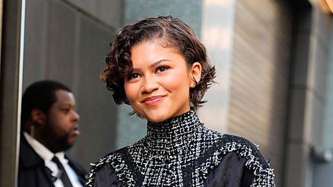 An image of actress Zendaya. She is smiling at the camera, wearing a black and white high-necked dress with her hair pulled back. 