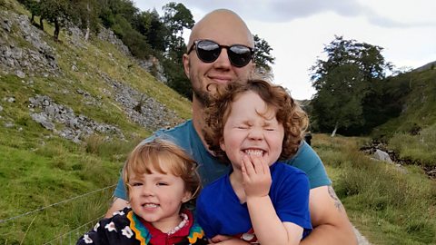 A dad stands holding his two children affectionately to camera