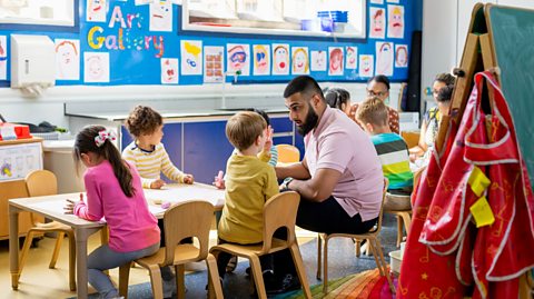 A nursery class with four children working at a table to the left, with one looking at their male teacher on the right. Another group is sat behind.