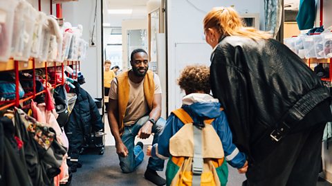 A male nursery teacher or assistant is looking towards a pupil and their mother, as if welcoming them to start the day.