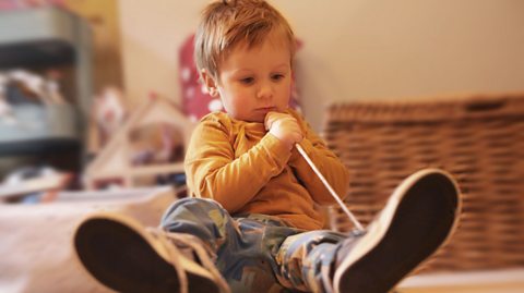 A close-up of a child who is tying their own shoe-lace, looking intently in the direction of their left shoe.
