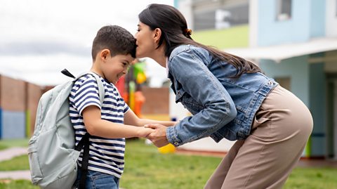 A mum stood with what appears to be her son; she is kissing him on the head, holding his hands, as if about to let him walk into a nursery or school setting.