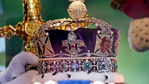 An image of the Imperial State Crown, an ornate golden crown with ermine trim, sat on a cushion. 