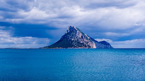 A landscape image shows a blue sea and a cloudy sky. In the distance is an island with Rocky Mountains. 
