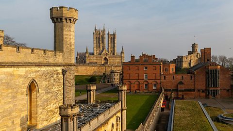 A landscape image of Lincoln Castle, a sprawling stone medieval castle with round turrets. In the background, Lincoln Cathedral can be seen.