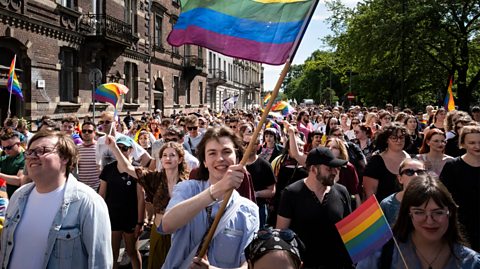 Participants march in the 22nd Kraków Equality Parade in Kraków, Poland. On a sunny day, the crowd walks towards the camera, with many smiling and waving Pride flags, which are rainbow flags with six horizontal stripes of different colours