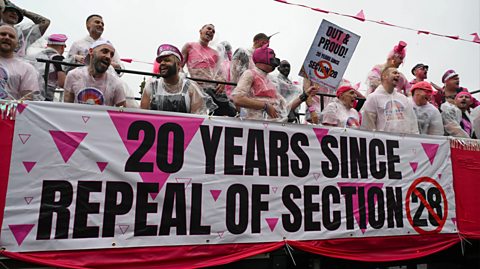 Brighton & Hove Pride 2023 - two decades on from the repeal of Section 28. A group stand on the top deck of an open top bus wearing white and pink. Over the side of the bust is a sign which reads ' 20 years since repeal of Section 28'