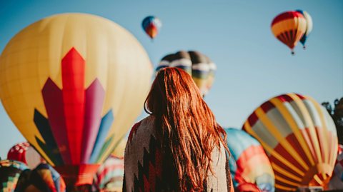 A woman with ginger hair and her shoulders wrapped in a blanket faces a number of hot air balloons taking flight. They vary in colour and are large balloons with baskets to carry passengers attached