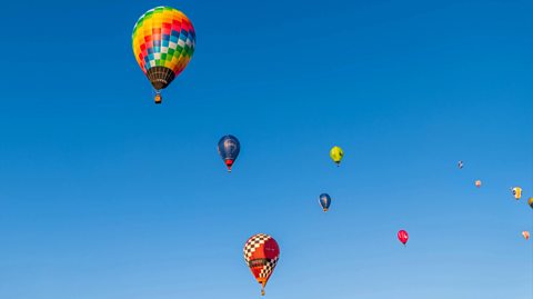 Multicoloured hot air balloons fly in the distance in the blue sky. They are large balloons with baskets to carry passengers attached