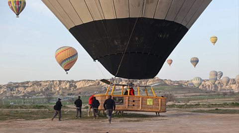 Before it takes flight, ground crew check a black and grey hot air balloon which has a wooden basket attached. In the distance against a blue sky, multicoloured hot air balloons are in flight