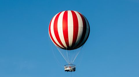 In the blue sky, a red and white striped hot air balloon is in flight. It is a large, spherical balloon with a basked attached to carry passengers