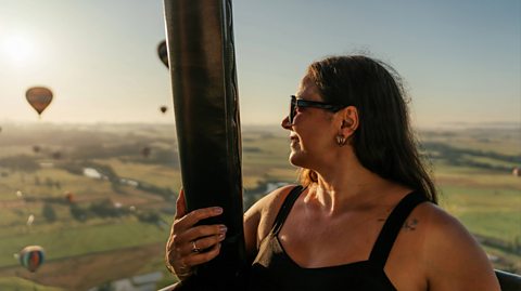 A woman with long brown hair, dark sunglasses and wearing a black strappy top smiles stands in a hot air balloon. She smiles looking out at the other hot air balloons in the distance. They are large balloons with baskets attached to carry passengers