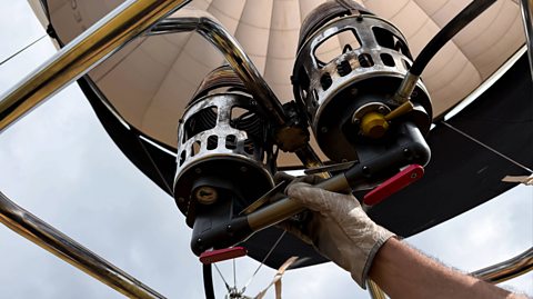 Hot air balloon flight preparation with dual burners. A hand wearing a glove holds a handle beneath the dual burners