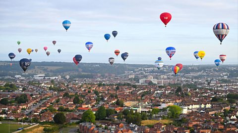 Over Bristol city centre, filled with green spaces and buildings, dozens of hot air balloons are pictured in flight. They all vary in colour and are large balloons with baskets attached to carry passengers