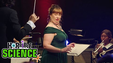 Image of Horrible Science character making a straining face while farting in an orchestra. She is wearing a green dress. The Horrible Science logo is in the bottom left.