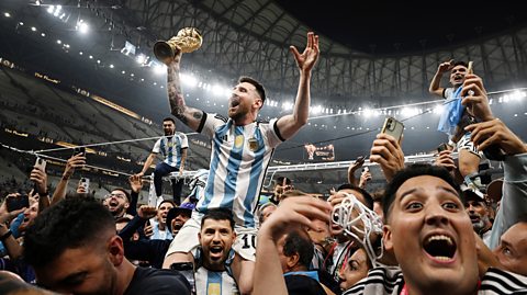 Lionel Messi celebrates with fans and team mates after Argentina wins the the FIFA World Cup 2022. He sits on a players shoulder holding a trophy in his white and blue striped football kit as the crowd cheers and celebrates around him