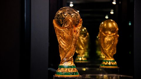 The FIFA World Cup trophy stands on display in a dark, mirrored room. The gold trophy depicts two human figures holding up the planet Earth.