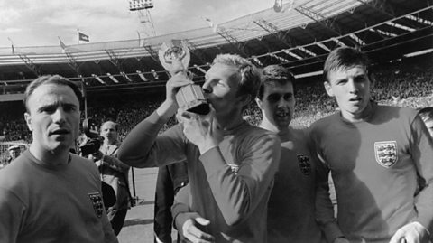 A black and white photo of players of the 1966 World Cup England team. Captain Bobby Moore kisses the World Cup trophy next to team-mates George Cohen, Geoff Hurst and Martin Peters join Moore in a victory lap. They all have long sleeved football tops on and behind them on the pitch is a camera operator, with the fans in the stands in the distance