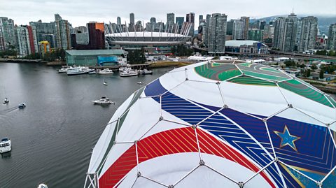 An aerial view of Science World during its transformation into "The Beautiful Dome," for the FIFA World Cup 2026 in Vancouver, British Columbia. Looking over the skyline and water, the dome is at the forefront of the image and looks like a white, blue, green and white footbal