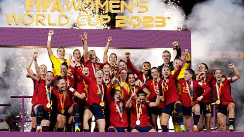 Spain lifts the trophy after winning the FIFA Women's World Cup in 2023. The team stand on a purple podium cheering in their red football kits
