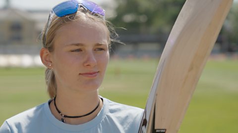 Tilly Corteen-Coleman, an England Women's cricketer, looks at a cricket bat whilst standing in a bright, sunny cricket ground.