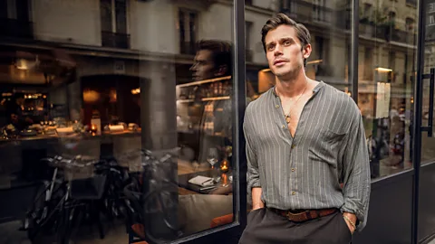 National Geographic/ Amy Browning Antoni Porowski wears a grey button down and stands in front of the glass windows of a restaurant (Credit: National Geographic/ Amy Browning)